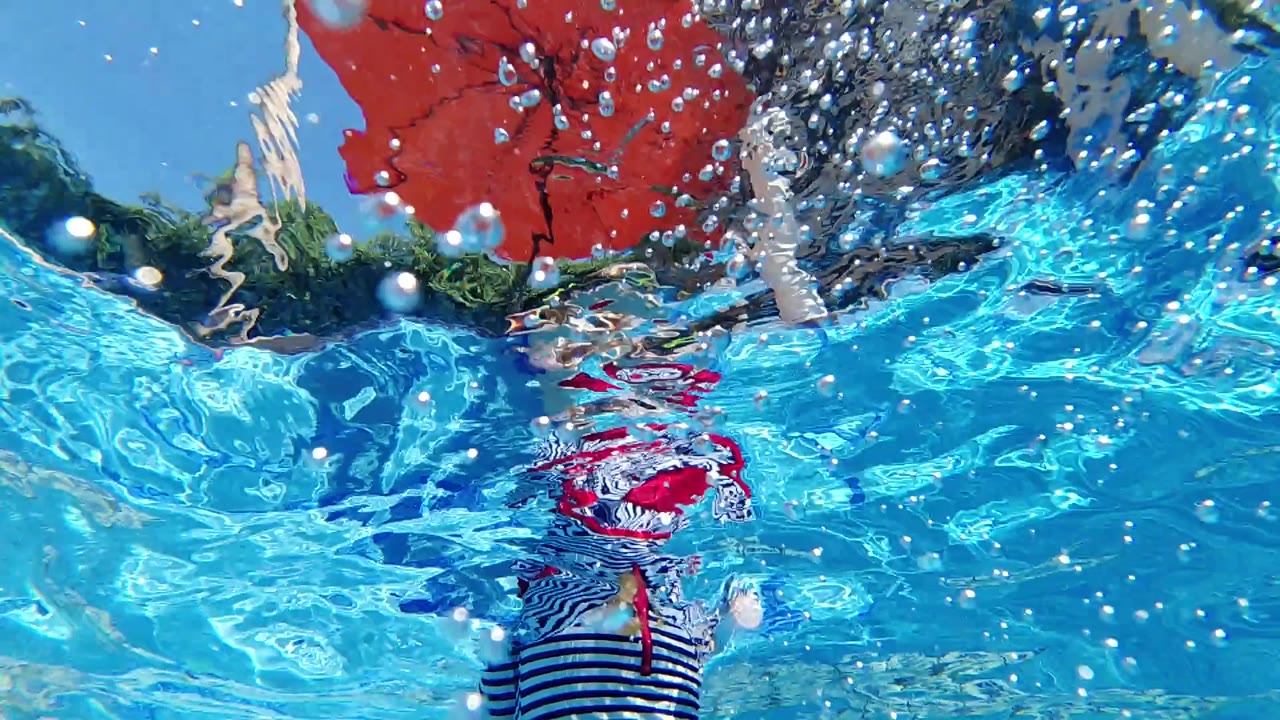 Blue pool water with bubbles, red umbrella, and striped swimwear distorted by ripples.