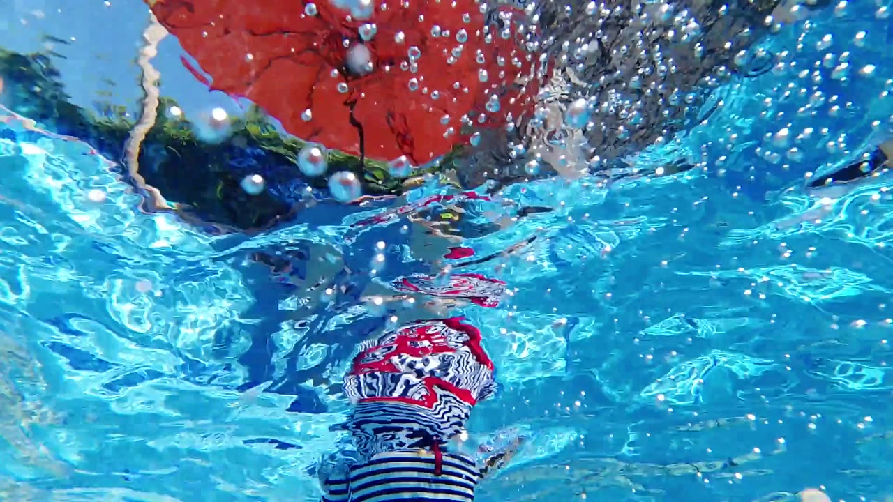 Rippling blue water covers the frame while the red umbrella and a person in red and striped swimwear appear warped through the surface.