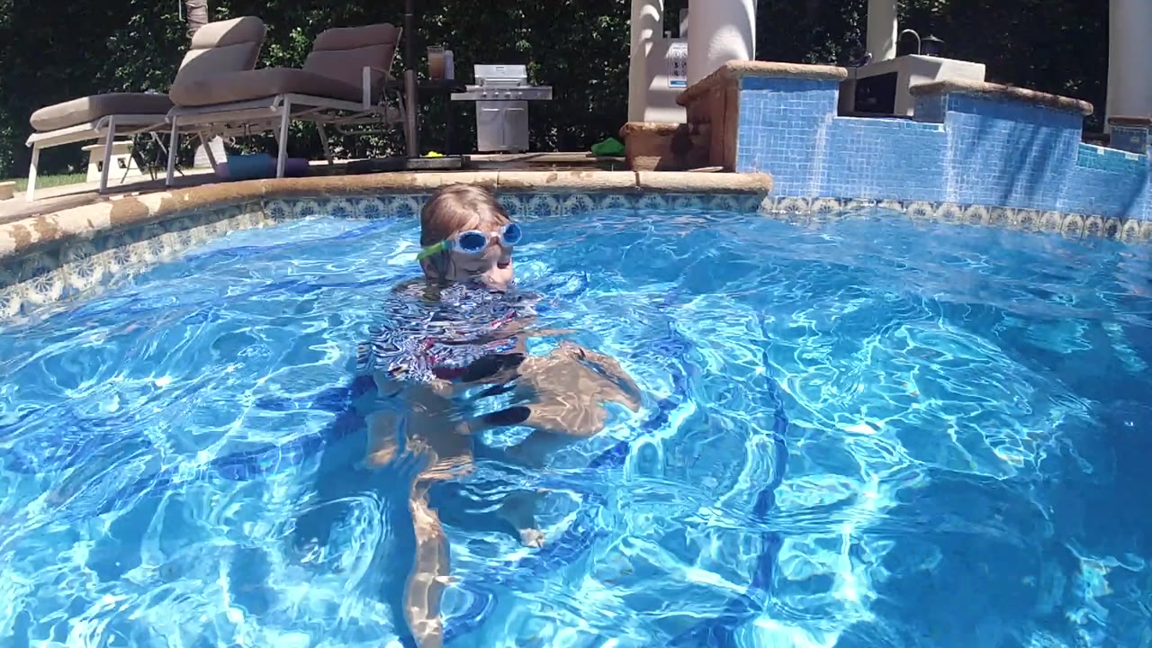 Young boy wearing blue goggles and a striped swim shirt floats near the center of a bright blue backyard pool in Cabo, with lounge chairs behind the tiled edge.