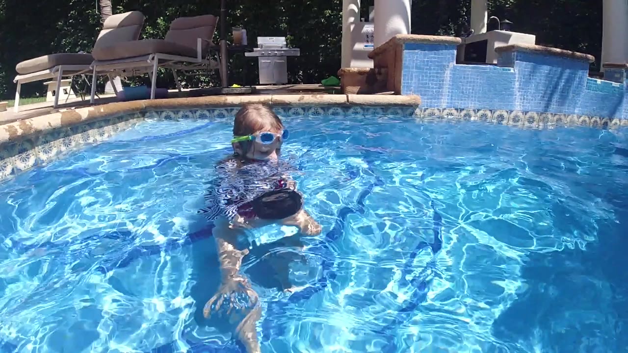 The child faces the camera from the pool, with one leg visible under the water and a tiled waterfall feature across the background in Cabo.