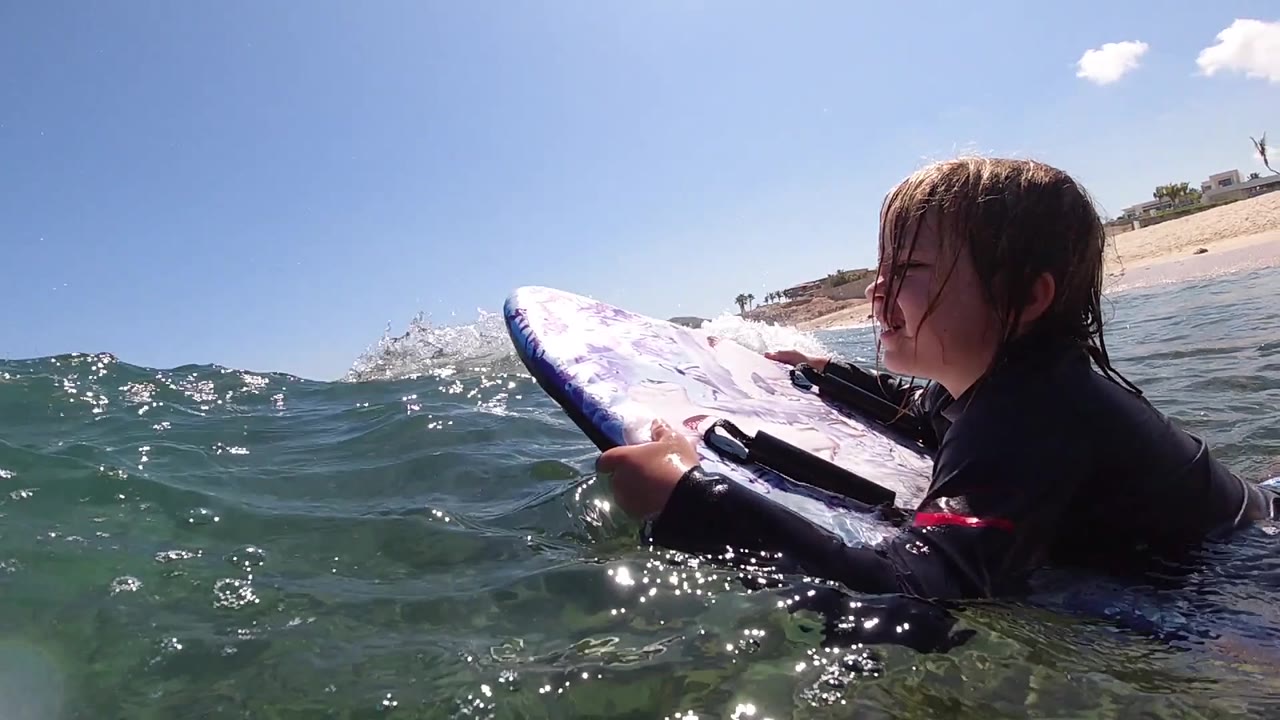 The surfer's profile is visible in Cabo, Baja California Sur, with sunlit droplets around the purple board and the sandy beach stretching behind.
