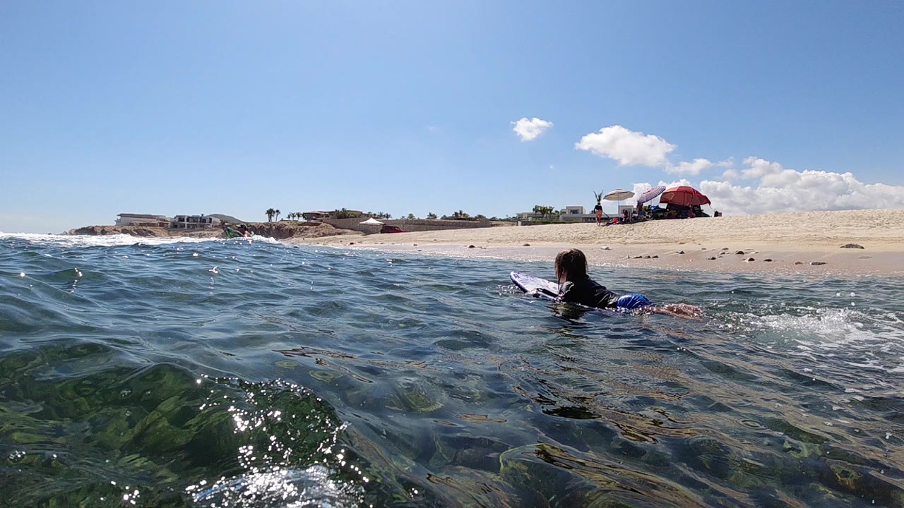 At 0:00, a surfer paddles on a light surfboard through blue-green water near the sandy shore in Cabo, Baja California Sur, with beach umbrellas and distant buildings behind.