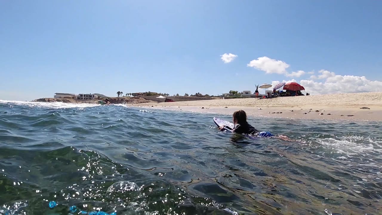 At 0:02, a small foreground wave partly lifts across the frame while the Cabo beach, red umbrella, white canopy, and paddling surfer remain visible in bright sun.