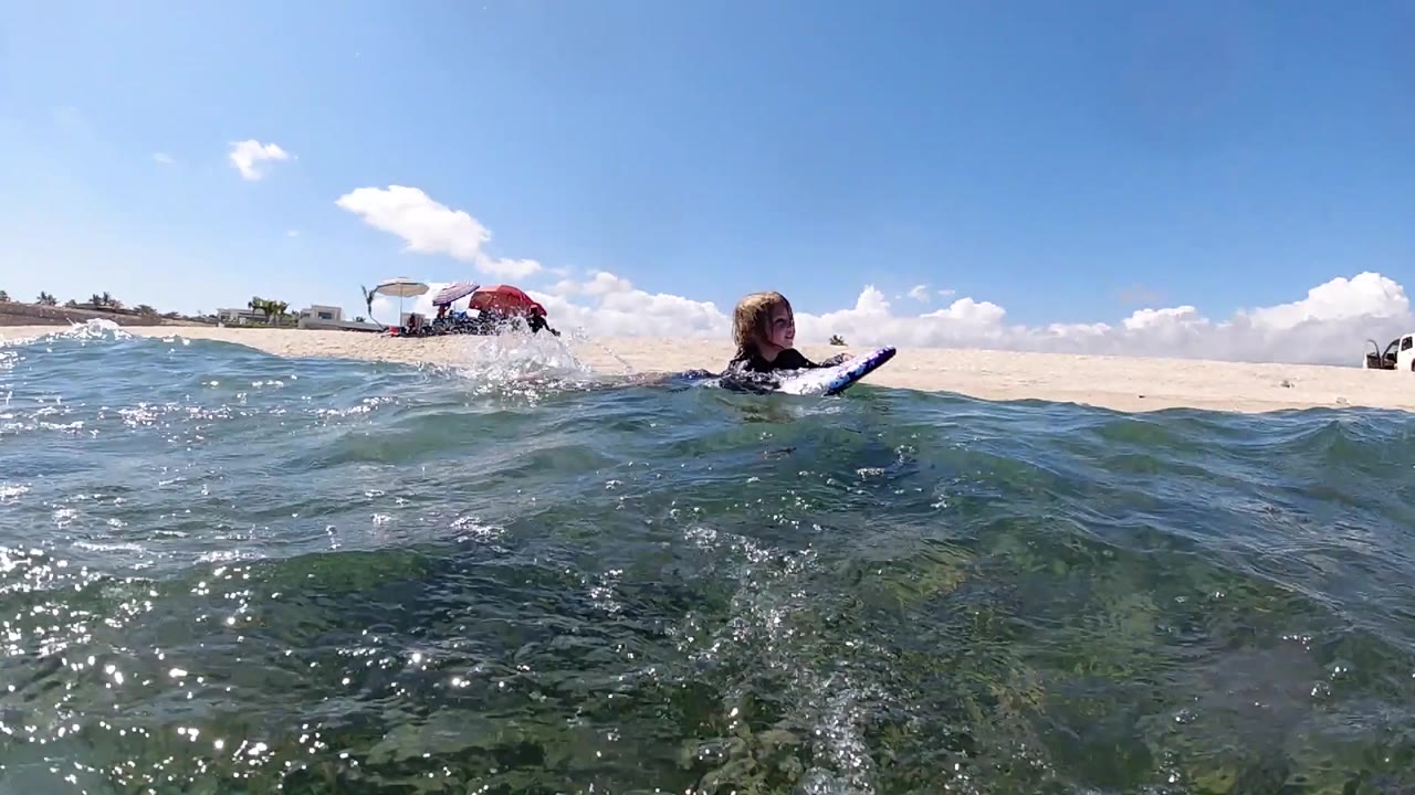 The surfer shifts right of center while choppy shallow water rises across the foreground and the sandy Cabo shoreline stays visible behind.