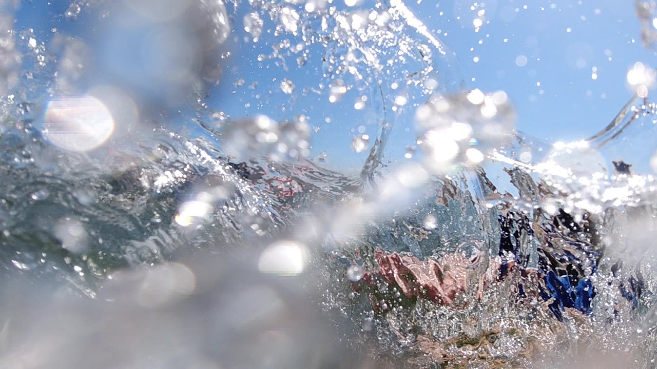 At water level in Cabo, a burst of surf spray partly obscures children bodyboarding under a bright blue sky.