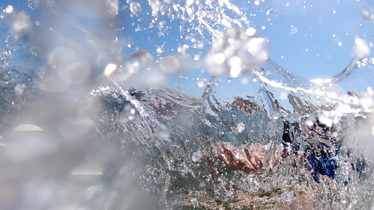 Spray and lens droplets cover a child paddling near another bodyboarder.