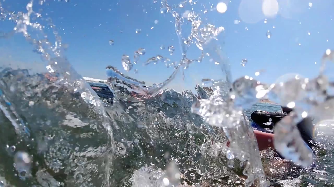 Breaking water fills the foreground as a child on a blue board and another on an orange board appear beyond the splash near the Cabo shoreline.