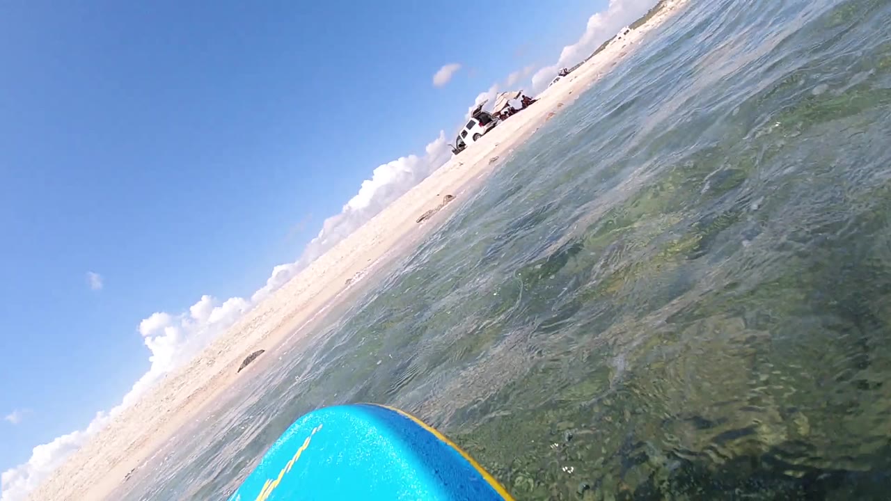 Tilted point-of-view shot from water surface level in Cabo, Baja California Sur, showing blue surfboard at lower left, sandy beach and shoreline at center, ocean water to the left, blue sky above; no people visible