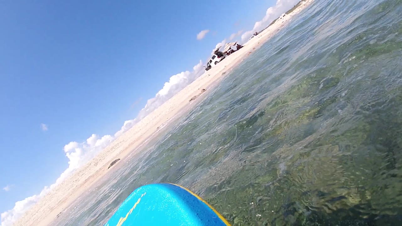 Continued tilted point-of-view from near water surface in Cabo, Baja California Sur, showing blue surfboard at lower center, sandy beach and ocean boundary in mid-frame, blue sky above; no people visible