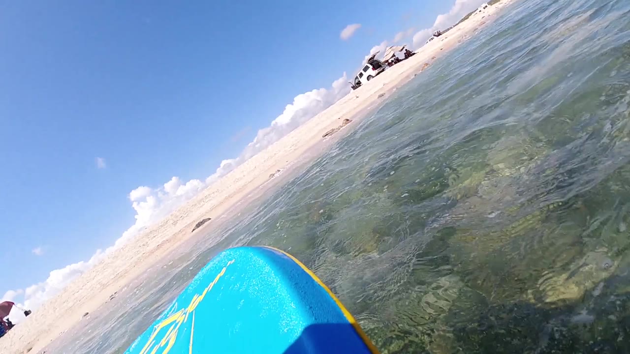 Tilted POV from water surface level in Cabo, Baja California Sur, blue surfboard at lower right, sandy beach and water boundary in center frame, sky above; no people visible