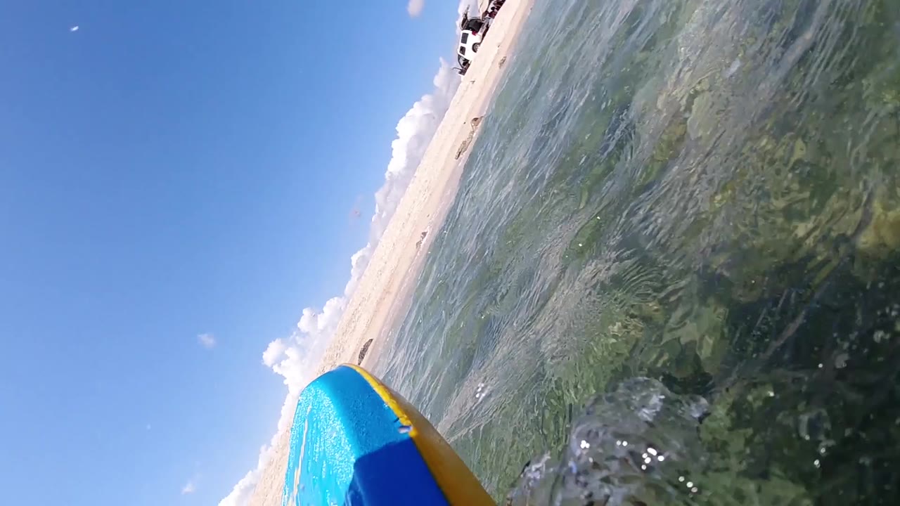 Tilted POV at water surface with blue surfboard at left and sandy beach across mid-frame