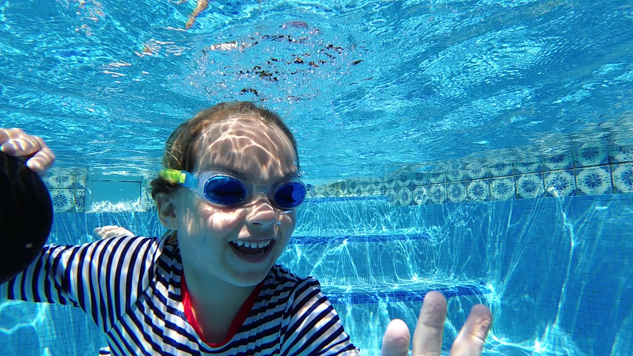 A young girl in blue goggles and a striped swim shirt smiles underwater in a clear pool in Cabo, Baja California Sur, with a hand near the foreground.