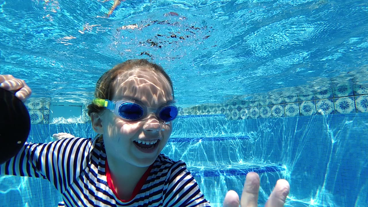 Smiling girl underwater with goggles and hand raised near camera.