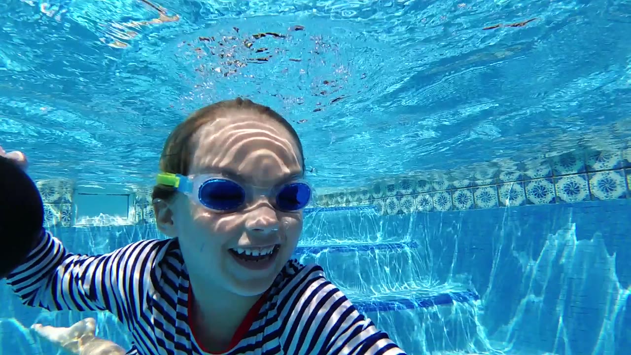 A laughing child in goggles fills the underwater foreground, with blue pool wall tile and sun patterns behind her in Cabo, Baja California Sur.