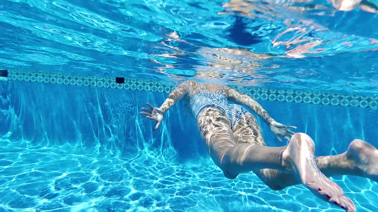 A swimmer glides farther across the underwater frame in Cabo, with blue pool water, caustic light patterns, and a tiled band along the wall.