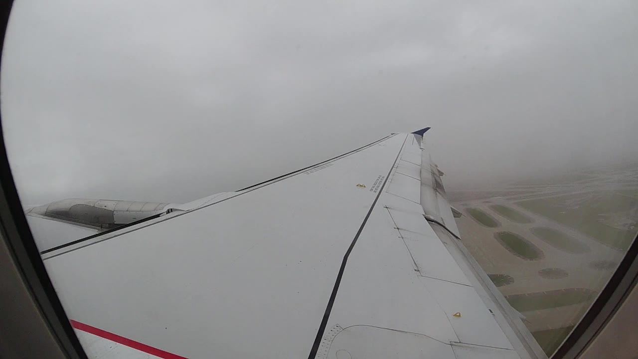 Opening frame from inside an aircraft cabin looking through the window at a white wing with a black leading edge and red livery stripe; gray overcast sky above, ground dimly visible through haze below, no visible people.