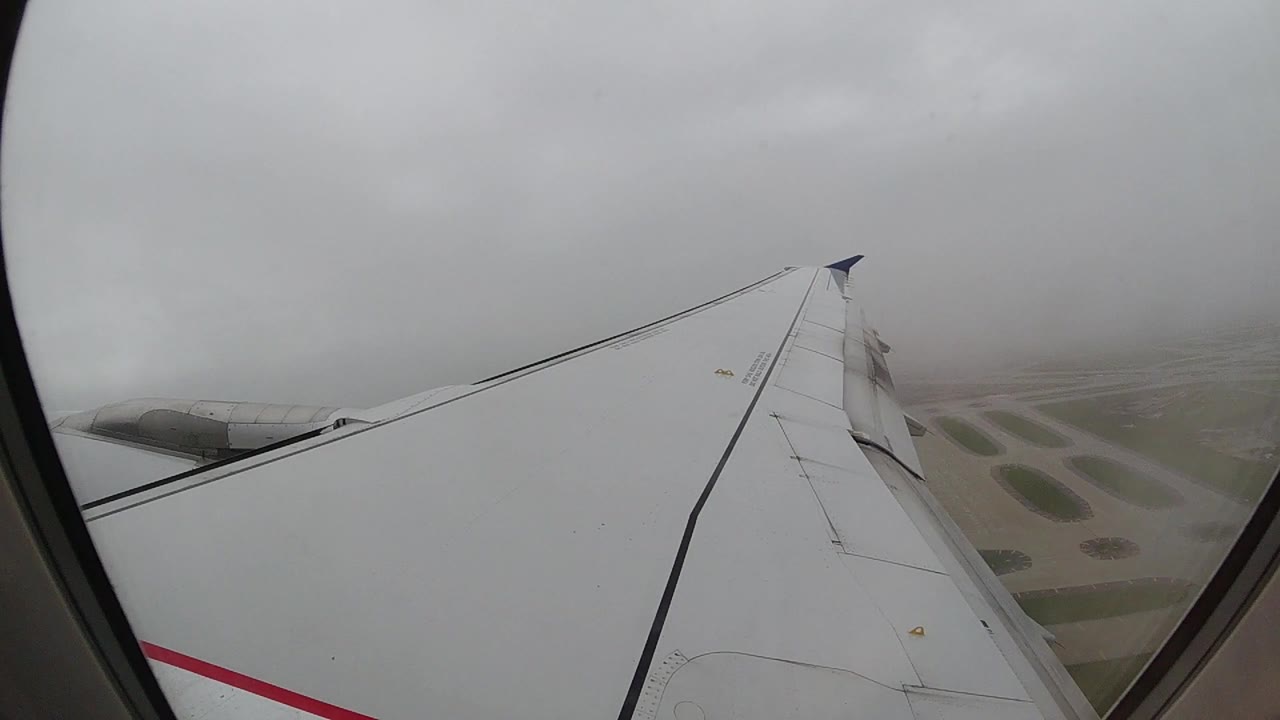 Airplane wing through cabin window, black leading edge prominent, flat terrain faintly visible below cloud layer.