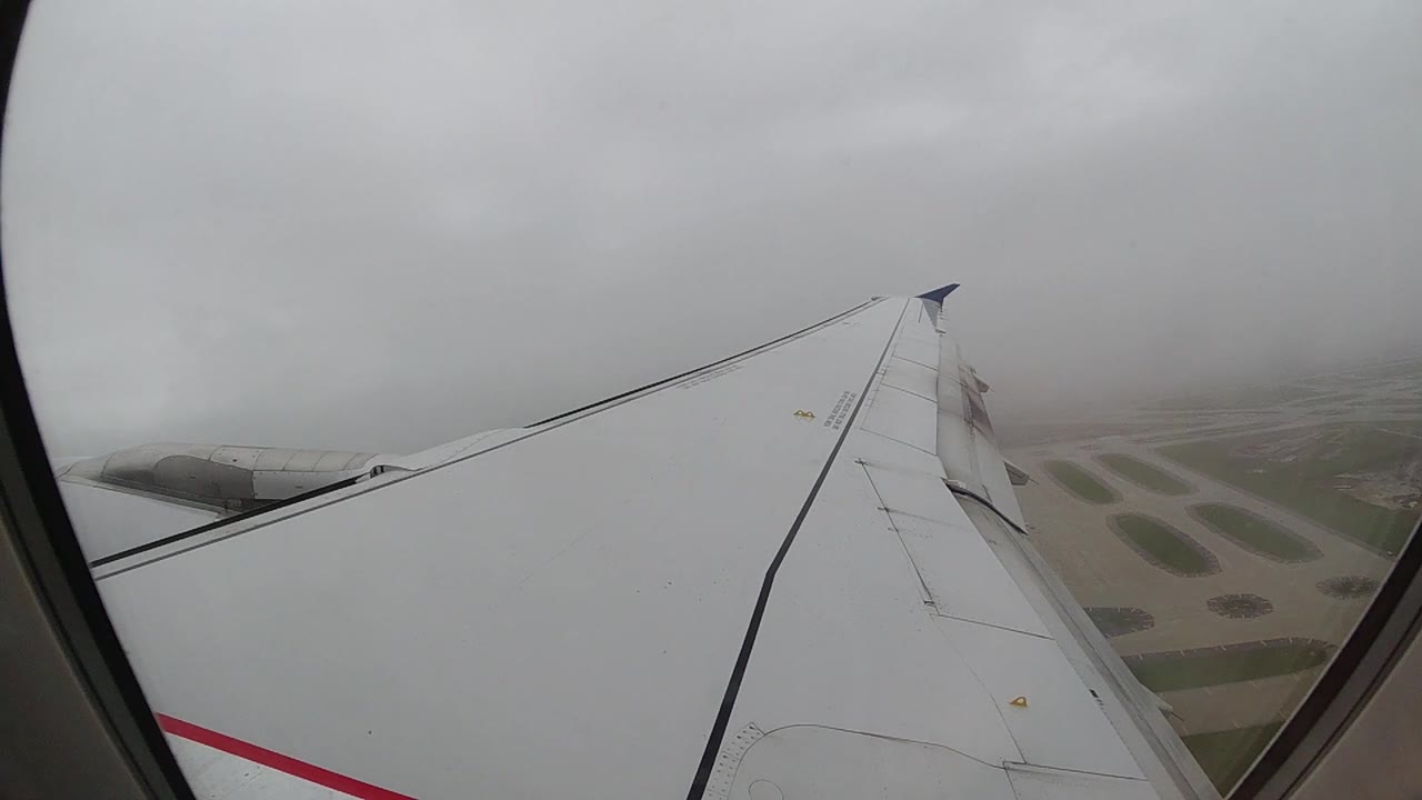White aircraft wing fills the center of the cabin window frame, black leading edge running diagonally toward the upper right, gray overcast sky above and cloud-obscured ground below, no visible people.