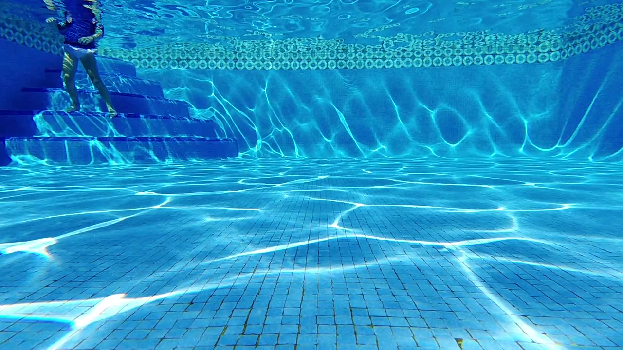 Underwater view in Cabo, Baja California Sur, Mexico, showing blue pool tiles, bright caustic light, left-side steps, and a swimmer's lower legs at the upper left.