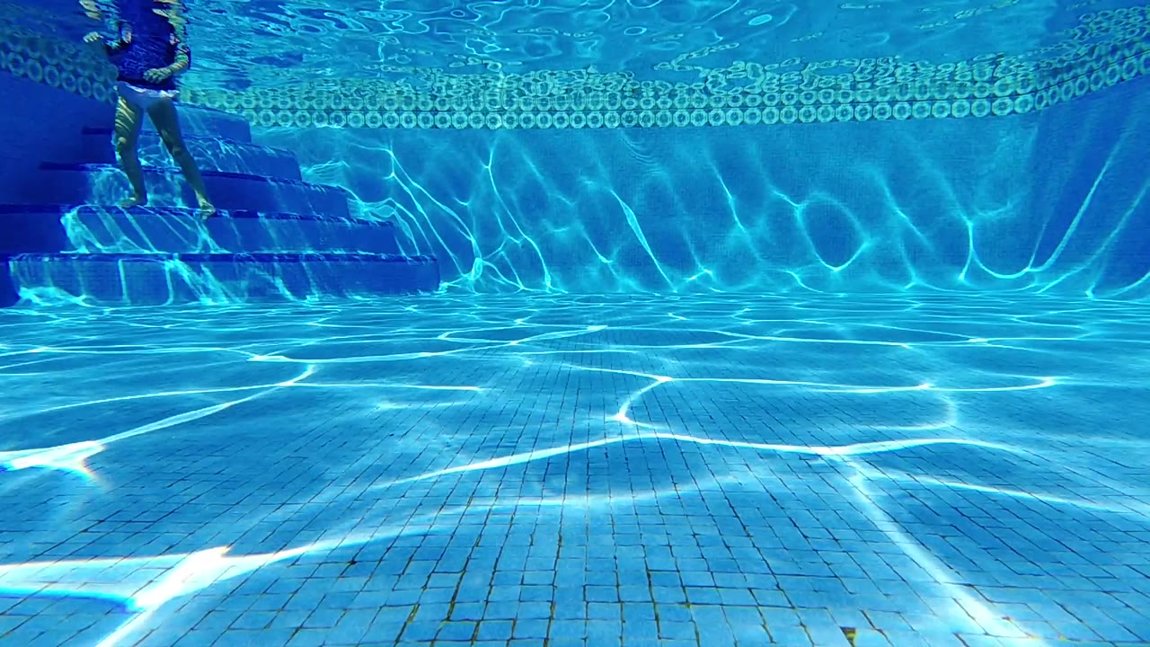 Blue tiled pool bottom with sun ripples and swimmer feet above left steps.