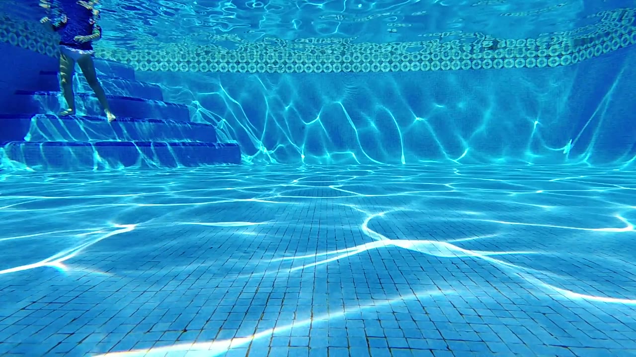 The Cabo pool frame shows strong white caustic lines crossing the blue-tiled floor while the swimmer's legs remain near the upper-left edge.
