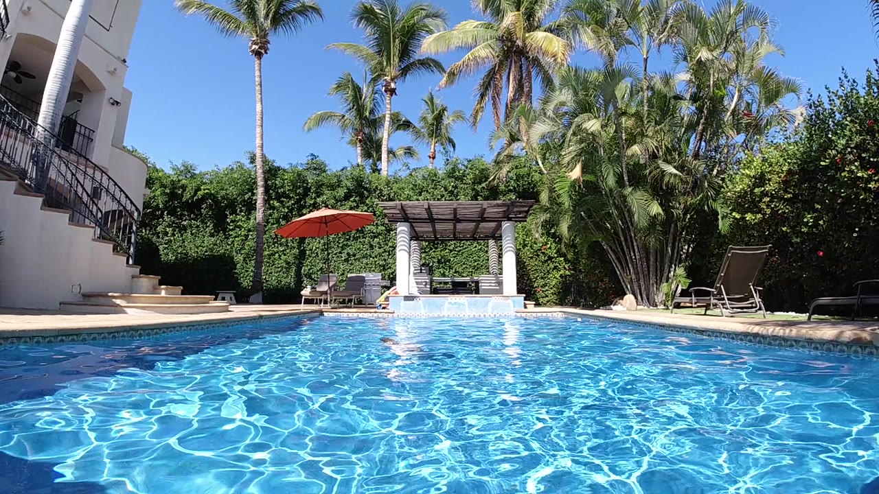 Blue pool water ripples toward a swimmer, villa stairs, palms, and gazebo.