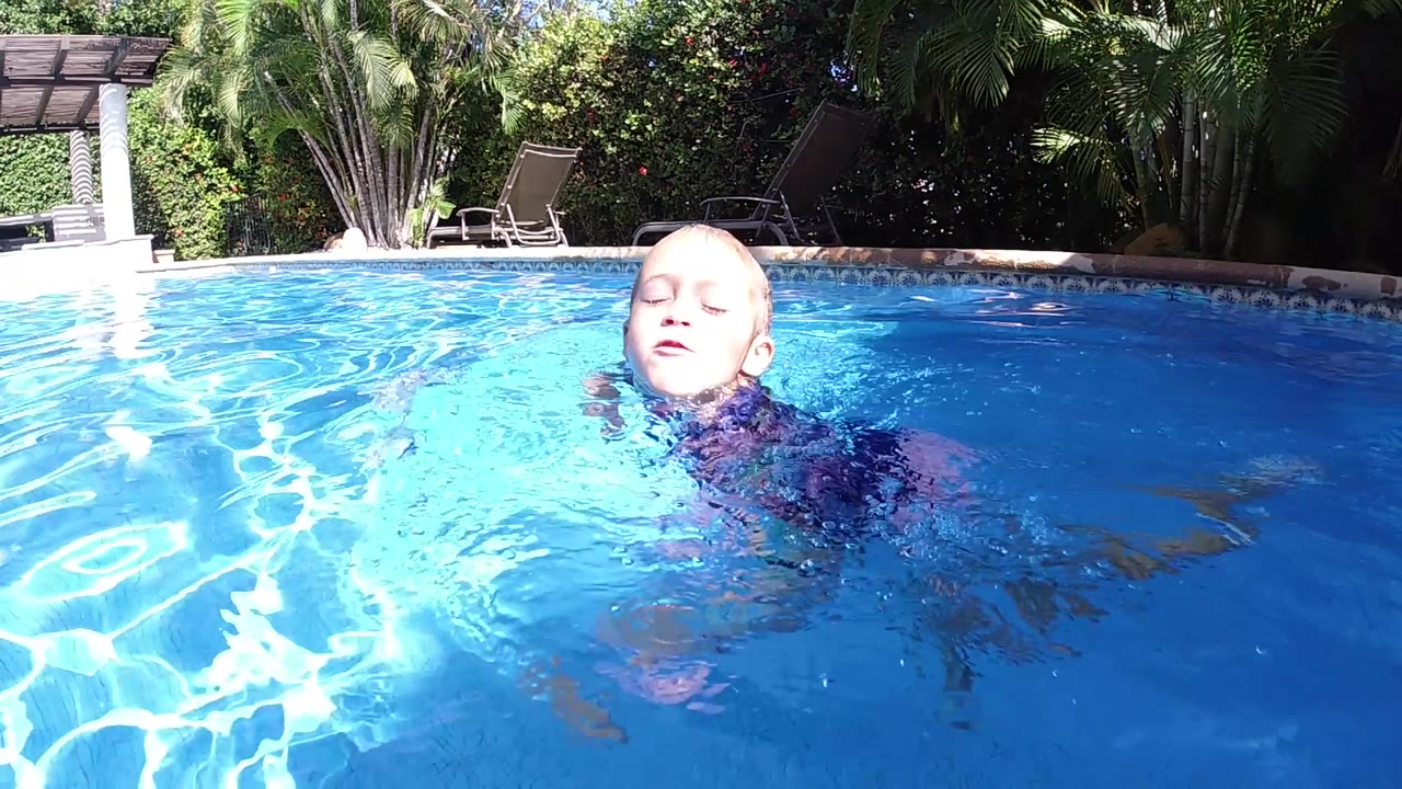 A blonde child turns her head slightly while swimming in the bright blue pool, with the shaded pergola structure and dense greenery visible behind her in Cabo, Baja California Sur.