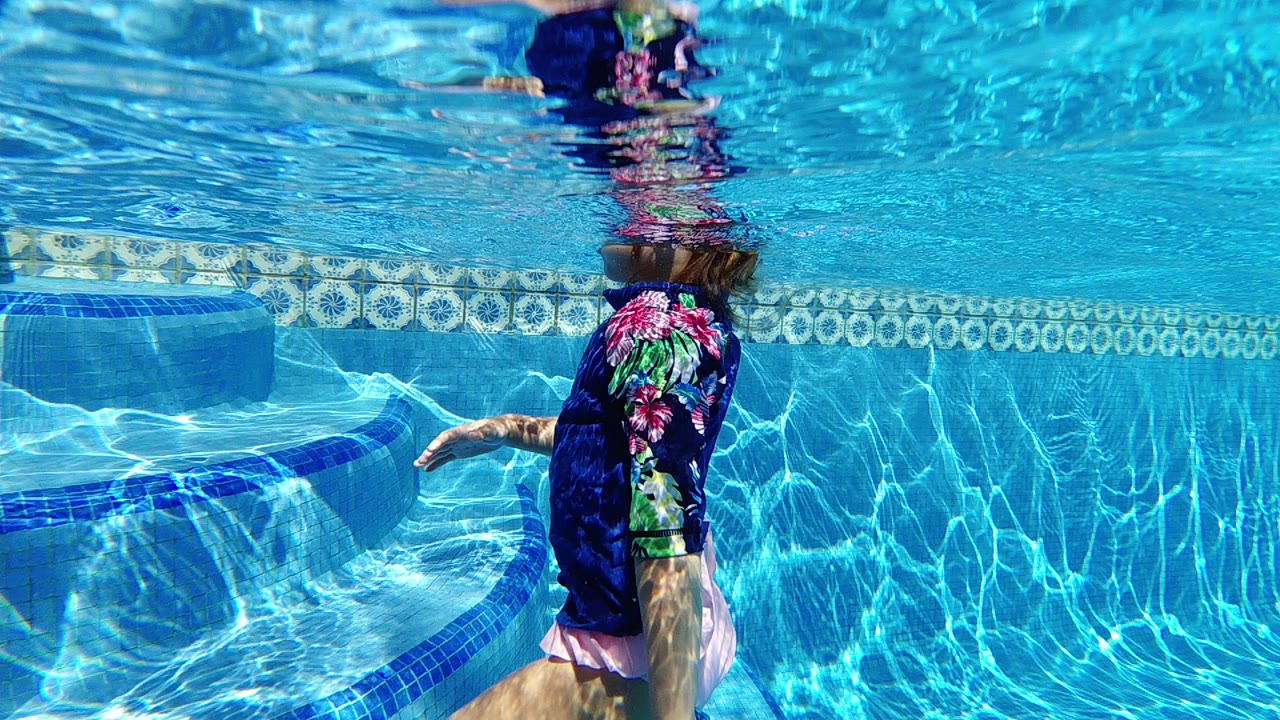 In Cabo, Baja California Sur, a woman in a dark rash guard is underwater beside curved blue pool steps, with her head near the surface and rippled light across the wall.