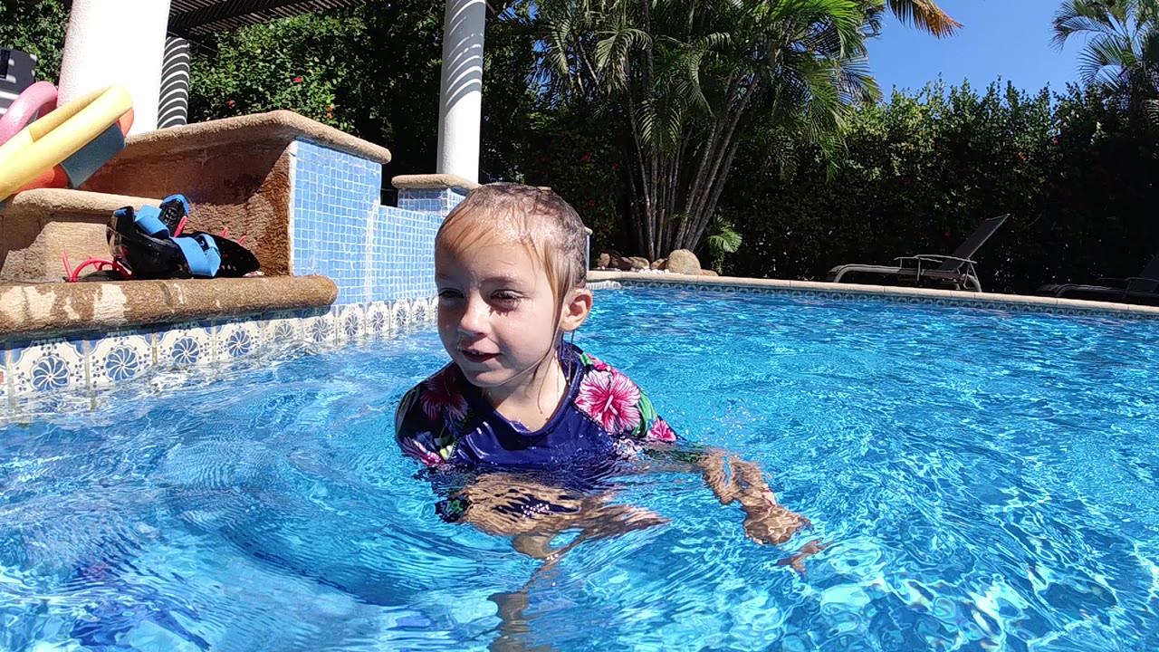 A young girl in a blue and pink swimsuit stands chest-deep in a bright pool in Cabo, Baja California Sur, with pool steps, noodles, palms, and lounge chairs behind her.