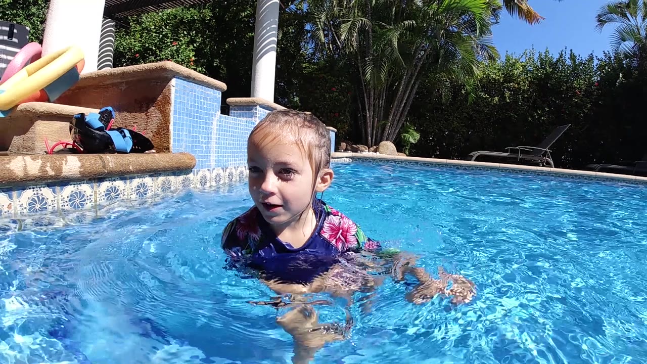 The child remains centered in the Cabo pool as her arm moves through the water, with bright blue reflections and shaded greenery behind the deck.