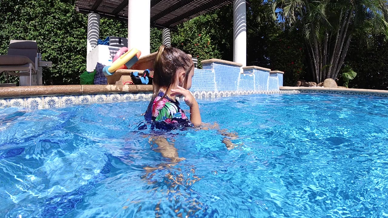 In Cabo, Baja California Sur, a young girl stands waist-deep in a bright blue swimming pool with her hand near her face and white patio pillars behind her.