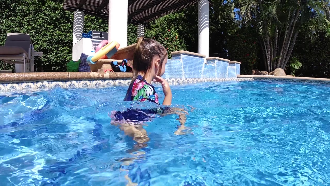 The girl faces across the swimming pool in Cabo while the sun lights the blue water, pool toys, white columns, and dense greenery in the background.