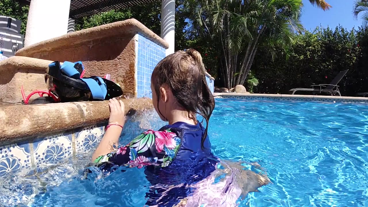 At 0:00 in Cabo, Baja California Sur, a young girl seen from behind holds the pool edge while bright water ripples around her and sandals sit on the ledge above.