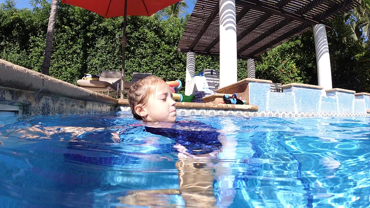 At the start of the Cabo pool clip, a young girl faces left while swimming through bright blue water, with white columns and a shaded patio behind her.