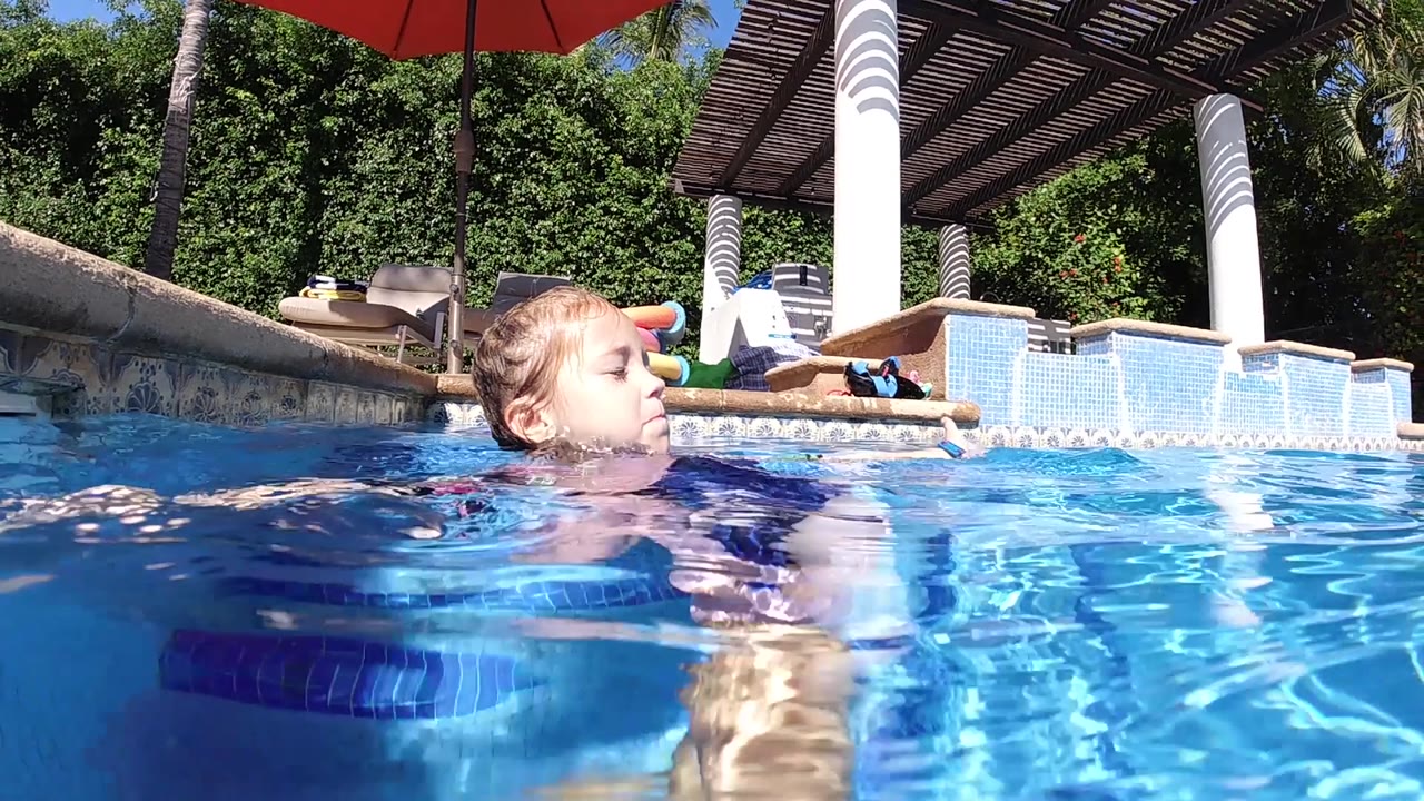 At 0:02, the child swims left to right through the frame, with stronger water reflections in front of her and lounge seating visible beyond the pool edge in Cabo.