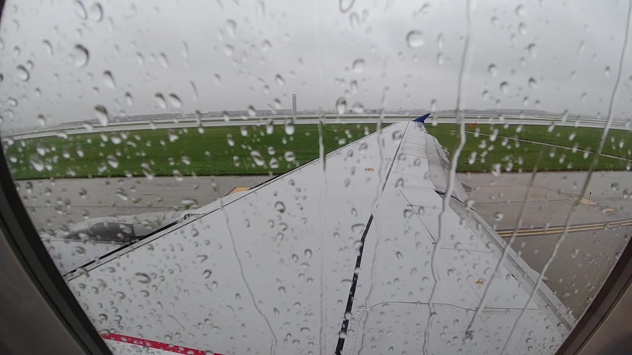 Heavy rain droplets on a passenger airplane window at a Chicago-area airport in 2019, with the white aircraft wing, grey tarmac, and green infield grass visible through the distorted glass; no people visible