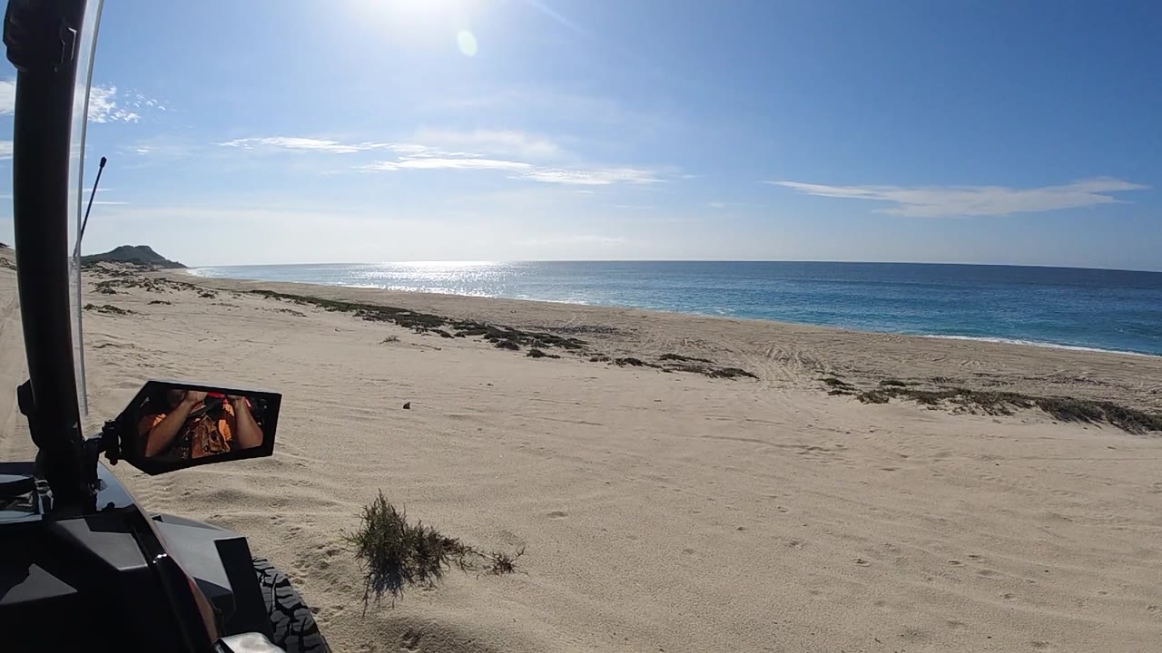 One second into the clip, the UTV continues along the beach in Baja California Sur; the roll cage and orange safety flag remain at left, pale sand fills the center, and the Pacific Ocean stretches to the right under a blue sky with no people visible.