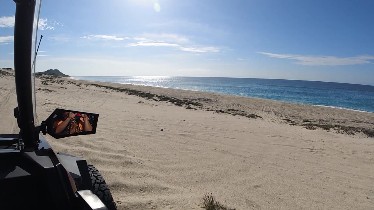 At 0:02, the off-road vehicle tracks along the Baja California Sur beach; the roll cage frames the left edge, pale sand stretches ahead, the Pacific Ocean is visible to the right, blue sky fills the upper frame, and no people are visible.