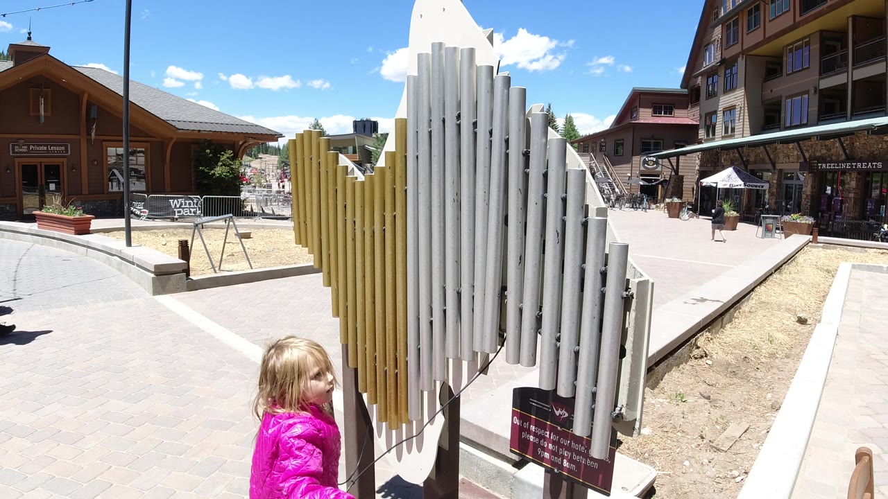The child in pink shifts position beside the white and yellow pipe sculpture, with sunlit paving, rustic buildings, and scattered pedestrians in the distance.