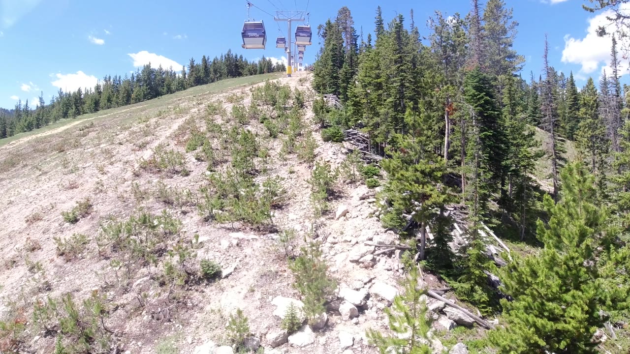 Aerial drone view looking forward along a Colorado ski run in summer, dry sandy terrain and pine forest flanking the corridor, chairlift cables crossing the upper frame edge, no visible people.