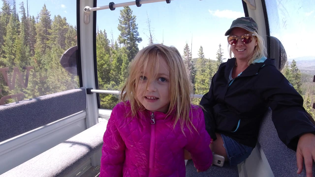 At the start, a blonde girl in a bright pink jacket faces the camera inside a Colorado mountain gondola while an adult woman sits behind her and pine trees fill the windows.