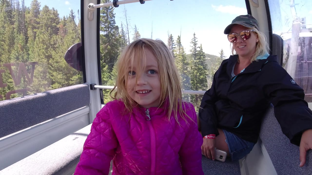 The girl smiles slightly inside the Colorado gondola, with the adult woman seated behind her, gray bench cushions around them, and pine trees outside the window.
