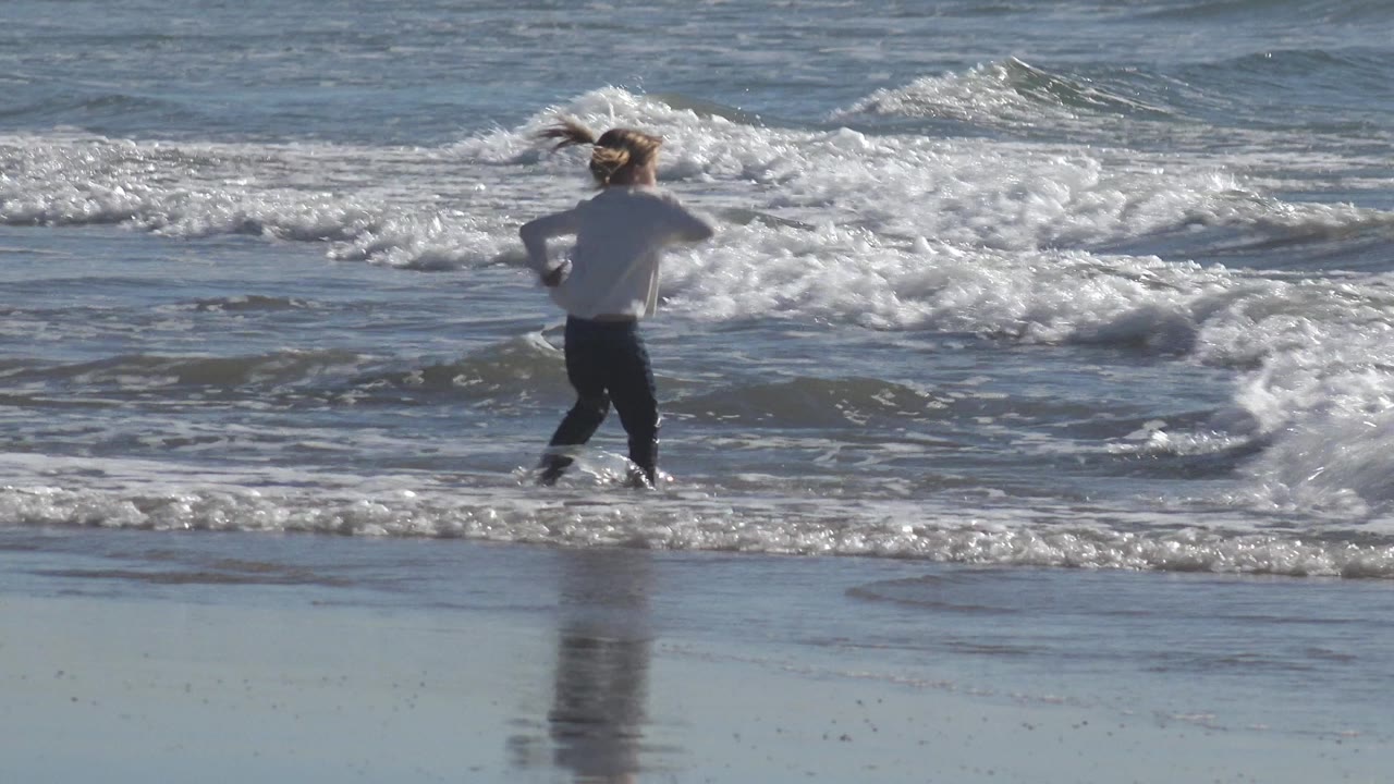 A child in a white shirt and dark pants runs toward shallow beach waves, with wet sand reflecting the figure and no adult visible in this cropped moment.
