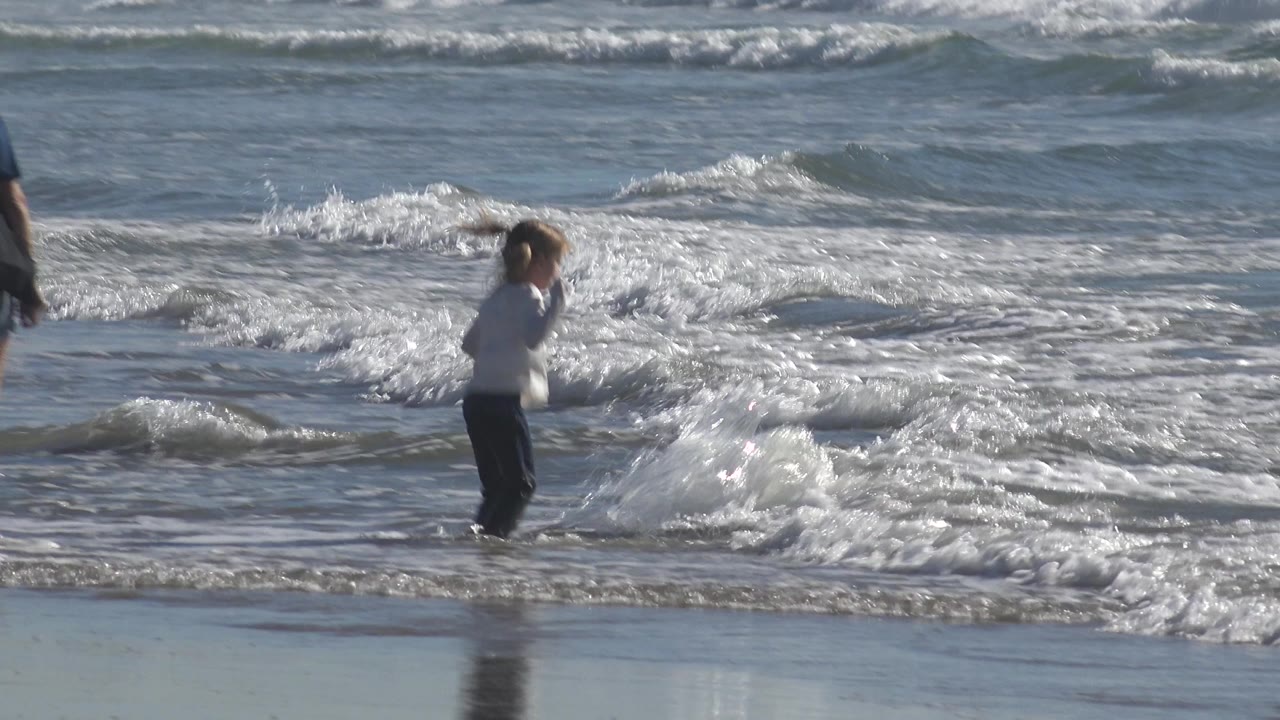 A child pauses at the edge of churning white foam, with sunlight catching the rippled ocean surface behind them.