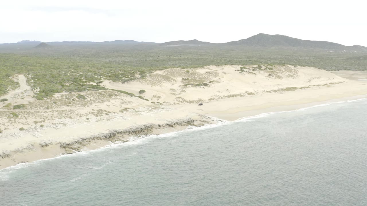 Aerial view at the start of the clip with gray-green ocean in the foreground, a pale sandy beach and dune ridge through the middle, scrub vegetation inland, distant hills, and no visible people.
