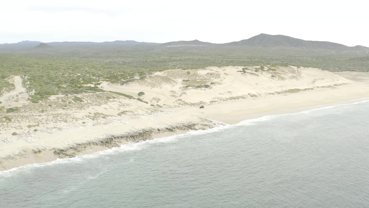 Aerial frame looks across calm gray water toward a sandy shoreline, pale dunes with scattered vegetation, a wide scrub-covered inland area, low hills on the horizon, and no visible people.