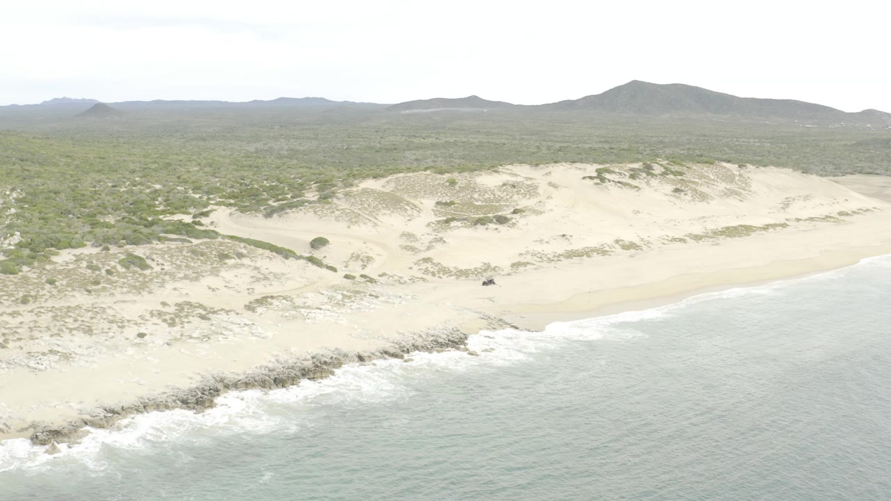 Aerial view with the rocky surf line lower in frame, pale dune ridges across the center, green scrubland behind them, low hills on the horizon, and no visible people.