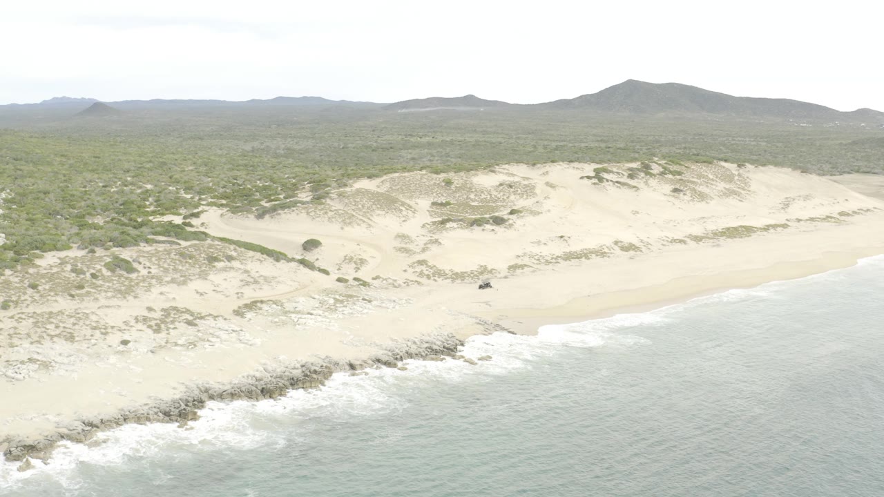 Wide aerial still showing ocean water at right, a pale sand beach below the dunes, scrub vegetation spreading inland, distant hills under a washed bright sky, and no visible people.