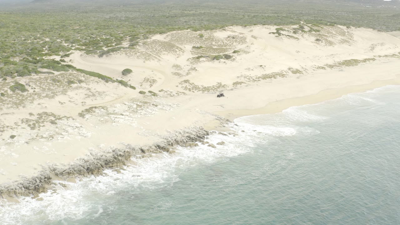 Aerial frame with the off-road vehicle slightly farther along the sand, backed by low dunes, scattered vegetation, rocks, and white waves rolling in, with no visible people.