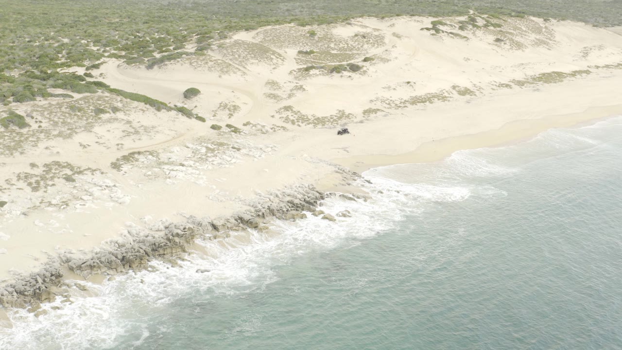High drone perspective looking across a sandy coastal curve where the small off-road vehicle sits near the dune side while surf breaks across rocks, with no visible people.
